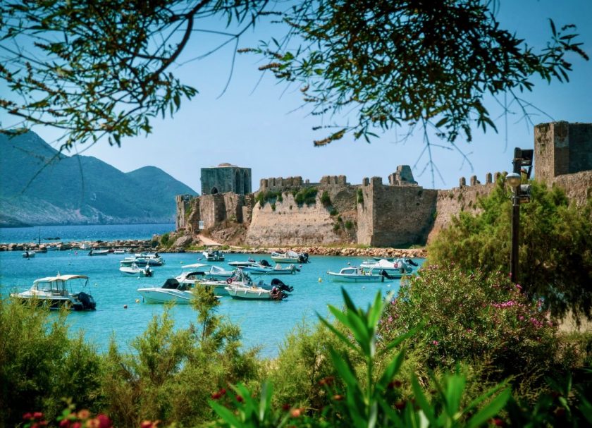Blick auf eine Festung am Wasser mit Booten und Bergen im Hintergrund.
